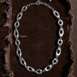 Op de afbeelding: Een zilverkleurige ketting met ovale schakels, elk met een centrale opening. De ketting wordt gepresenteerd tegen een donkerbruine, getextureerde achtergrond. De schakels zijn gelijkmatig verdeeld, waardoor een symmetrisch ontwerp ontstaat.