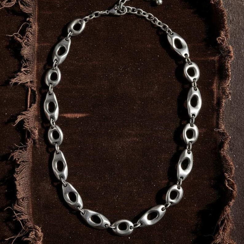 Silver chain necklace with oval sculptural links arranged on dark brown textured fabric. Each link features an elongated open center, creating a bold, modern industrial look. Contemporary unisex jewelry photographed in natural light with a raw, artisanal mood.