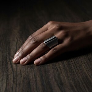 Hand wearing a wide silver statement ring with an open geometric design, resting on a dark wooden surface under soft, moody lighting.