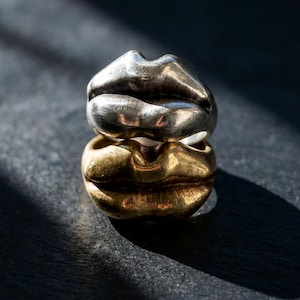 Könnte beinhalten: Zwei übereinander gestapelte Ringe in Lippenform, einer silberfarben, der andere goldfarben. Sie liegen auf einem dunklen Untergrund, mit einem Schattenwurf durch das Sonnenlicht. Einzigartiger Schmuck.