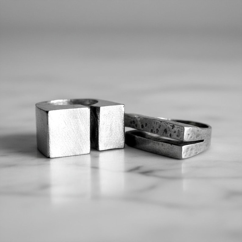 Two silver sculptural rings placed on a marble surface — one with a clean cubic open form, the other a textured split-bar design. Both feature a matte industrial finish and minimalist geometry. Contemporary artisan jewelry by Rannka.