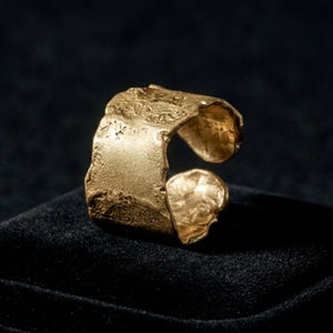 Puede incluir: Un anillo ancho y abierto de color dorado con una superficie texturizada e irregular y acabado mate. La banda presenta un aspecto rugoso y orgánico con bordes irregulares y un centro hueco, mostrando un color dorado metálico en toda la pieza.