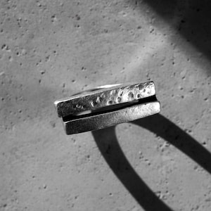 Close-up of a geometric silver ring with a hammered bar design casting a shadow on textured concrete. The raw metal surface and minimalist shape highlight its modern, architectural character. Unisex handcrafted jewelry by Rannka.