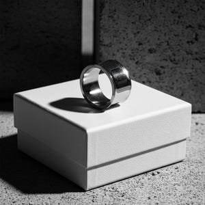 Wide stainless steel ring with a smooth reflective finish, displayed on a white jewelry box under directional light. The clean lines and contrast between metal and shadow highlight its bold, architectural form. Modern handcrafted design by Rannka.