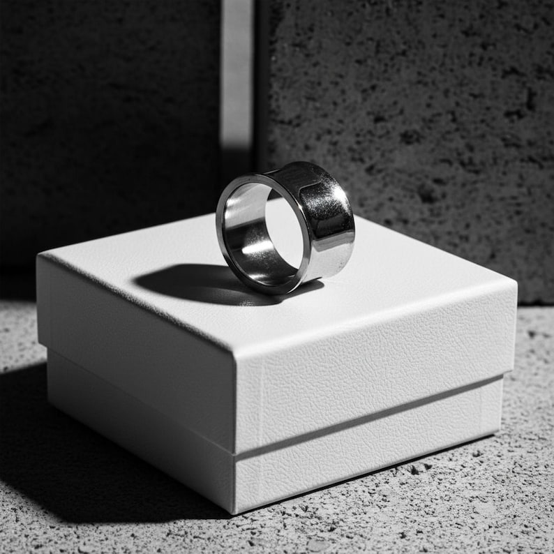 Wide stainless steel ring with a smooth reflective finish, displayed on a white jewelry box under directional light. The clean lines and contrast between metal and shadow highlight its bold, architectural form. Modern handcrafted design by Rannka.
