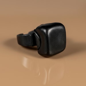 Puede incluir: Un anillo negro con forma cuadrada y bordes redondeados. El anillo está hecho de un material liso y mate. El anillo está sobre un fondo neutro. Se aprecia la banda del anillo.