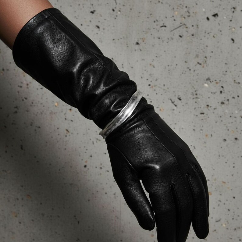 Silver cuff bracelet styled over long black leather gloves. The matte and metallic textures play off each other, giving the piece a sharp, modern edge. The shot focuses on contrast and material tension—clean, minimal, and industrial in tone.