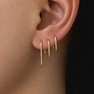 May include: Three gold-toned U-shaped earrings of varying lengths are inserted into three separate piercings along the earlobe. Each earring features a smooth, minimalist bar design that hangs vertically from the ear, creating a layered effect.