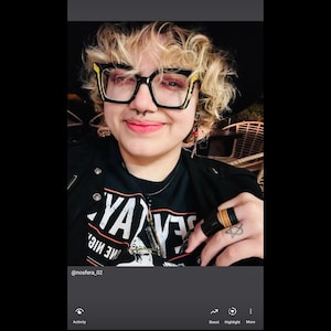 May include: A person wearing black-framed glasses with yellow accents, a black t-shirt with white text, and a black ring with a gold band. The person has a nose ring and a silver necklace. The image includes the text "YA" and "ME MIC."