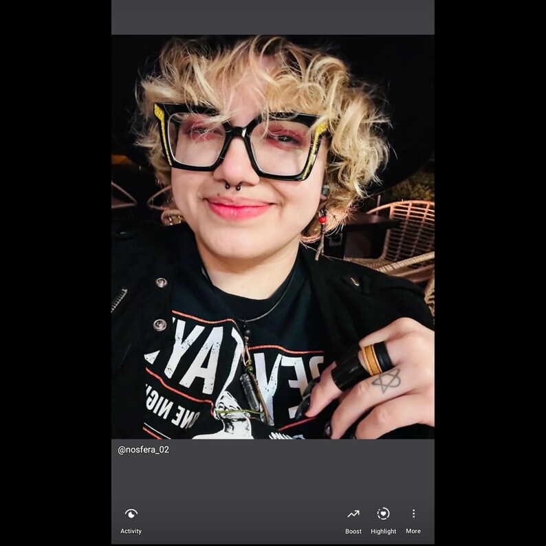 May include: A person wearing black-framed glasses with yellow accents, a black t-shirt with white text, and a black ring with a gold band. The person has a nose ring and a silver necklace. The image includes the text "YA" and "ME MIC."