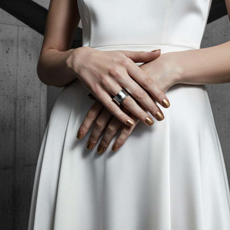 Close-up of hands resting over a white satin dress, featuring a polished stainless steel wide band ring. The mirror finish reflects soft light, blending industrial precision with minimalist elegance. Contemporary jewelry design by Rannka.