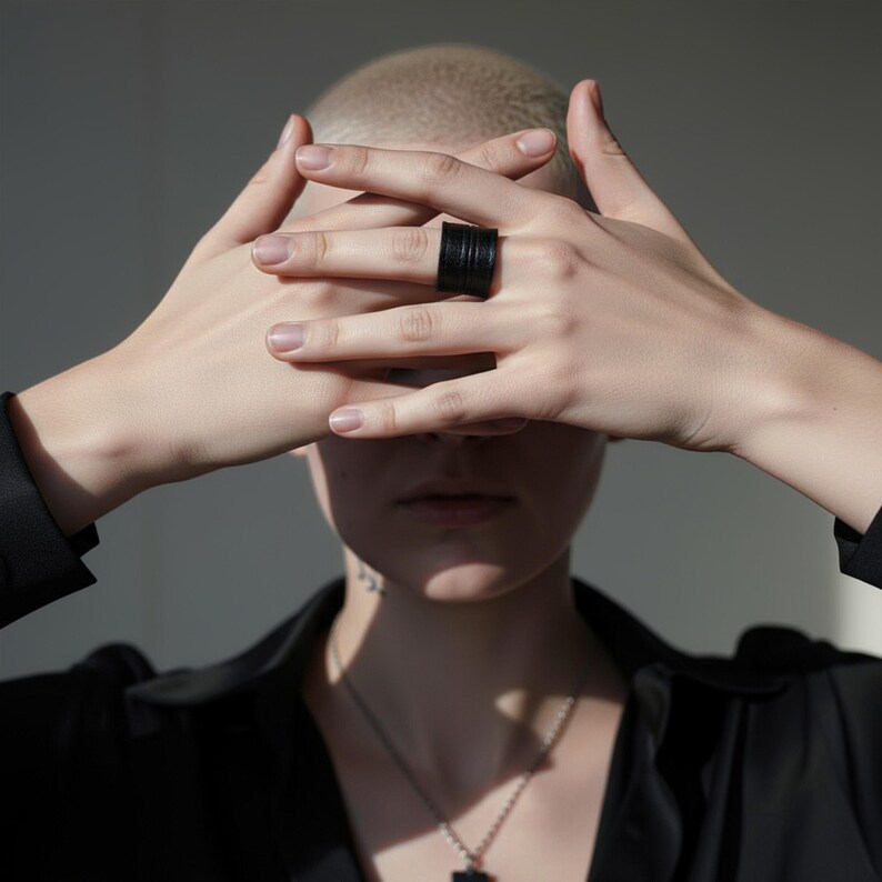 Person with hands crossed in front of the face wearing a single black vegan leather wrap ring. Clean editorial portrait with soft directional light and neutral background, highlighting the matte leather texture and sculptural band. Minimal contemporary cruelty-free ring by Rannka, styled with a quiet, modern, architectural mood.
