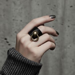 Hand wearing black vegan leather ring with antique brass sphere and loop accent. Dark ribbed sleeve and concrete background create a raw minimalist aesthetic, emphasizing the textured material and bold sculptural design of the statement ring.
