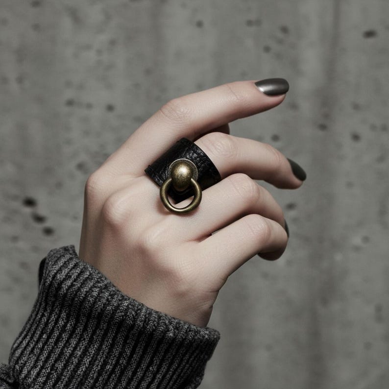Hand wearing black vegan leather ring with antique brass sphere and loop accent. Dark ribbed sleeve and concrete background create a raw minimalist aesthetic, emphasizing the textured material and bold sculptural design of the statement ring.