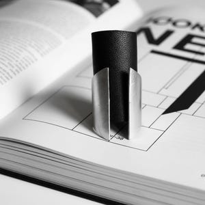 Black vegan leather and brushed silver ring photographed on an open design magazine. The clean geometric shapes of the page echo the ring’s angular metal edges, creating a visual dialogue between jewelry and modern graphic layout.