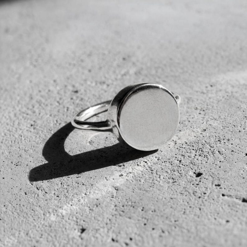 925 sterling silver round signet ring, clean flat surface with soft brushed finish, bright light casting a crisp shadow on textured stone background, minimal and precise with a cool, refined tone