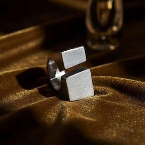 Bold open silver ring with squared ends, resting on golden velvet under warm light. The matte finish and clean geometry emphasize its sculptural form. Contemporary minimalist jewelry with industrial edge. Rannka design.