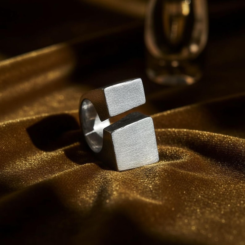 Bold open silver ring with squared ends, resting on golden velvet under warm light. The matte finish and clean geometry emphasize its sculptural form. Contemporary minimalist jewelry with industrial edge. Rannka design.