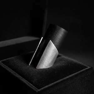Close-up of a wide black vegan leather and matte steel ring displayed on a dark textured box. The piece features angular, sculpted edges with a clean modern silhouette. Shot in low-key lighting to highlight contrast, material texture, and refined minimalist design.