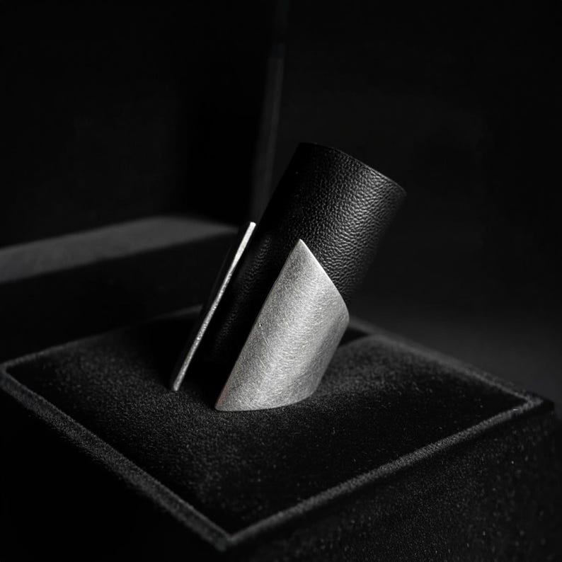 Close-up of a wide black vegan leather and matte steel ring displayed on a dark textured box. The piece features angular, sculpted edges with a clean modern silhouette. Shot in low-key lighting to highlight contrast, material texture, and refined minimalist design.