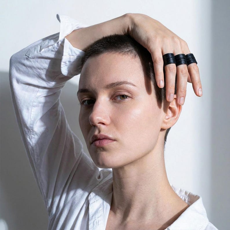 Portrait of a person wearing three black vegan leather wrap rings stacked across the fingers, hand resting on the head. Clean editorial image with soft natural light and a white background, highlighting the layered band construction and matte leather texture. Minimal sculptural ring cuffs with an architectural feel, styled with a white shirt for contrast. Contemporary cruelty-free jewelry by Rannka, shown for modern styling, everyday wear, and design-focused gifting.