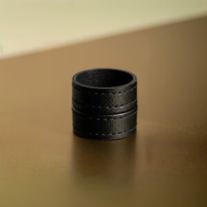 Single black vegan leather wrap ring standing upright on a warm gold surface, photographed in soft natural light. Minimal sculptural ring with layered band detail and matte grain texture, shown off-hand to highlight form, thickness, and material. Contemporary cruelty-free jewelry by Rannka with a refined, architectural mood.