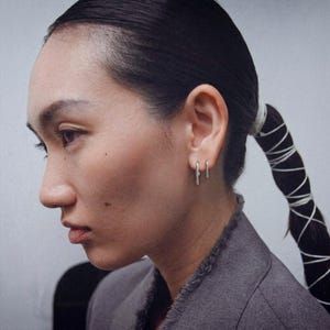 Sterling silver slip earrings in small and medium lengths worn as a minimal ear stack, clean bar drop earrings on ear, modern sculptural jewelry styling, subtle asymmetric look, everyday silver earrings, side profile portrait with slicked-back hair and tailored jacket, refined industrial aesthetic, Rannka style