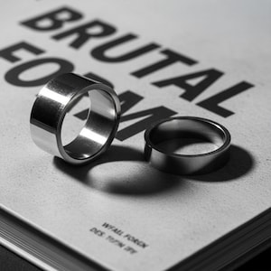 Two stainless steel band rings in different widths placed on a book cover reading “Brutal Form.” The rings cast clean shadows under direct light, highlighting their precision-cut edges and reflective surfaces. Modern industrial jewelry by Rannka with brutalist influence.