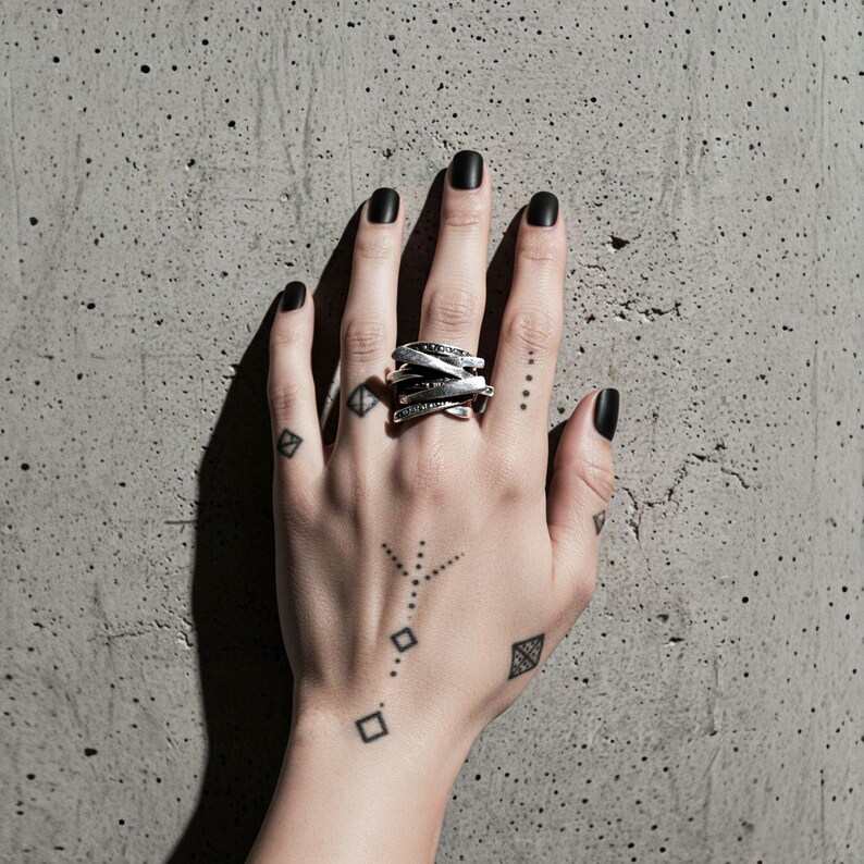 Tattooed hand with black matte nails wearing an oxidized layered silver ring featuring overlapping bands and dotted texture. The hand rests on a rough concrete surface, highlighting the ring’s industrial, geometric, and artisan design. Modern statement jewelry by Rannka.