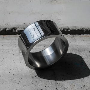 High-polished steel band ring with mirror reflection, resting on textured concrete in natural light. The piece features clean industrial lines and precision edges, blending modern craftsmanship with minimalist design. Contemporary statement jewelry by Rannka.