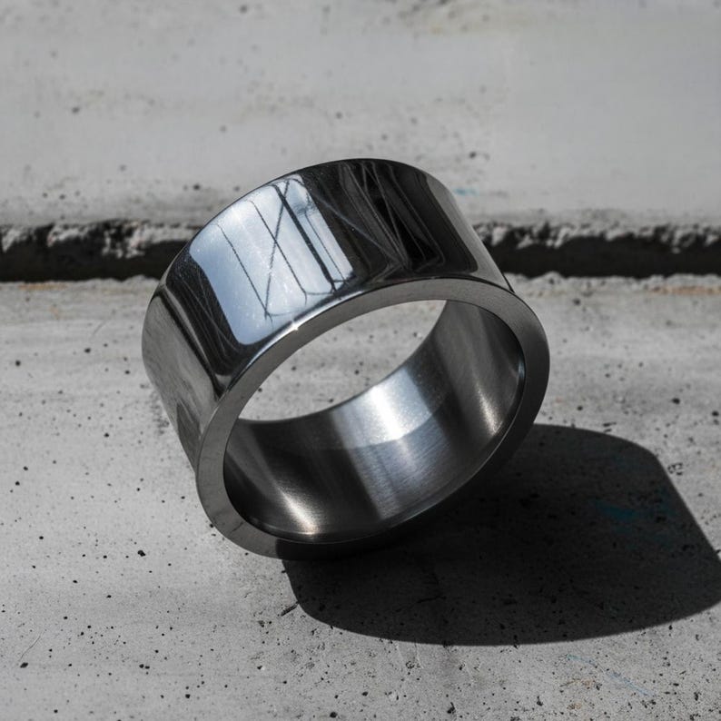 High-polished steel band ring with mirror reflection, resting on textured concrete in natural light. The piece features clean industrial lines and precision edges, blending modern craftsmanship with minimalist design. Contemporary statement jewelry by Rannka.