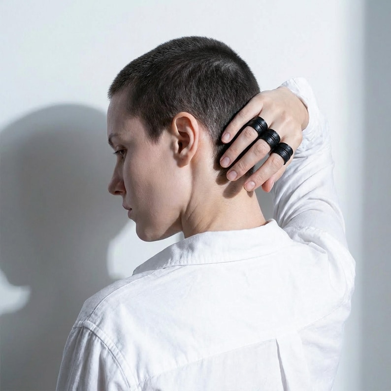 Side profile portrait of a person wearing three black vegan leather wrap rings stacked across the fingers, hand resting at the back of the head. Sculptural leather ring cuffs with layered band construction and matte grain texture are clearly visible, highlighting scale and fit when worn. Contemporary cruelty-free jewelry by Rannka, styled for modern ring stacking, everyday wear, and design-led gifting.