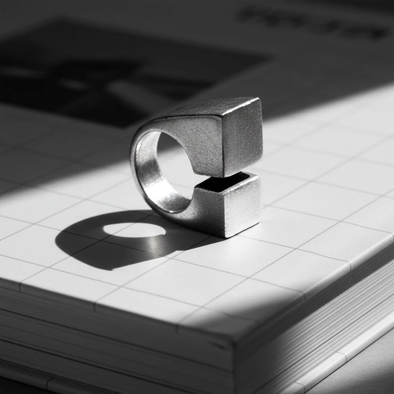 Architectural silver ring with squared open ends, photographed in black and white on a grid-patterned book. Clean lines, matte texture, and sharp light create a modern industrial mood. Minimalist sculptural jewelry by Rannka.