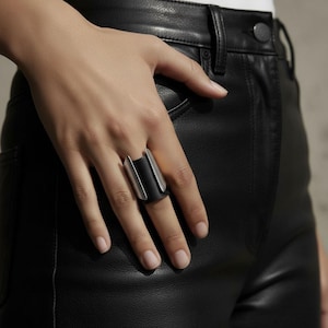 Hand wearing a wide black vegan leather and steel ring, styled with black leather pants. The ring’s sculpted metal edges contrast with the matte leather band, creating a modern industrial look. Minimalist styling, natural light, and clean composition emphasize contemporary design.