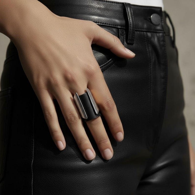 Hand wearing a wide black vegan leather and steel ring, styled with black leather pants. The ring’s sculpted metal edges contrast with the matte leather band, creating a modern industrial look. Minimalist styling, natural light, and clean composition emphasize contemporary design.