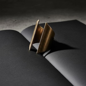 Sculptural open gold ring standing upright on folded black paper, matte brushed metal with an architectural form, minimalist product composition, modern jewelry design with sharp lines and negative space, Rannka statement ring.