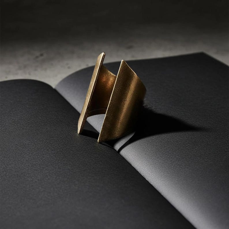Sculptural open gold ring standing upright on folded black paper, matte brushed metal with an architectural form, minimalist product composition, modern jewelry design with sharp lines and negative space, Rannka statement ring.