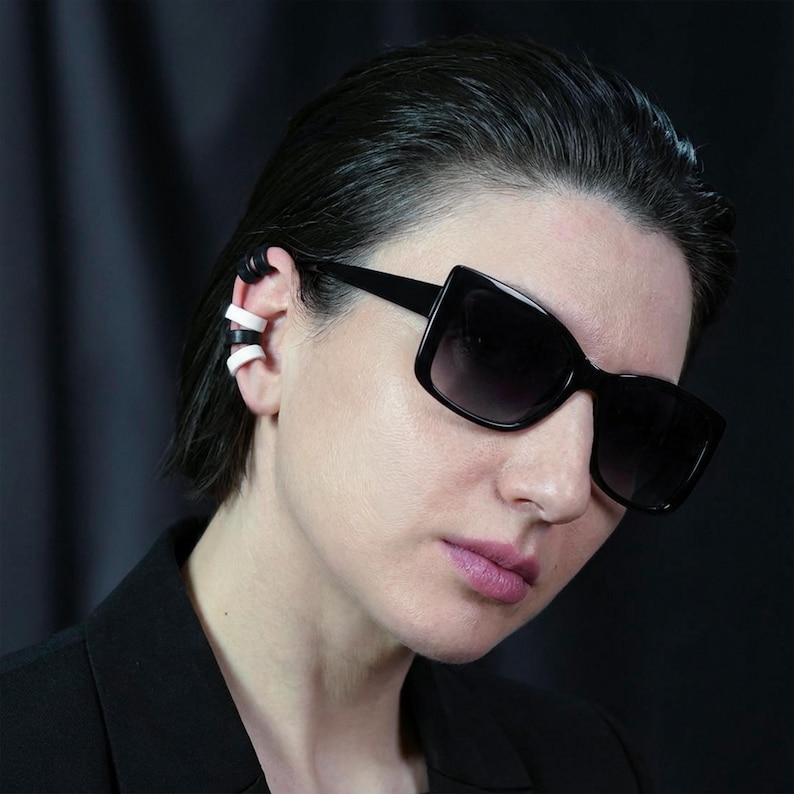 Black and white ear cuff stack worn along the ear without piercings, modern ear jewelry styled with sunglasses, sculptural ear wraps creating a layered ear look, minimalist statement ear accessory in dark studio lighting, contemporary no hole ear adornment by Rannka.