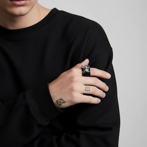 Person wearing black vegan leather ring with polished silver sphere and loop detail. Hand with geometric tattoos resting on black sleeve, minimalist styling with neutral background highlighting the modern unisex jewelry design.