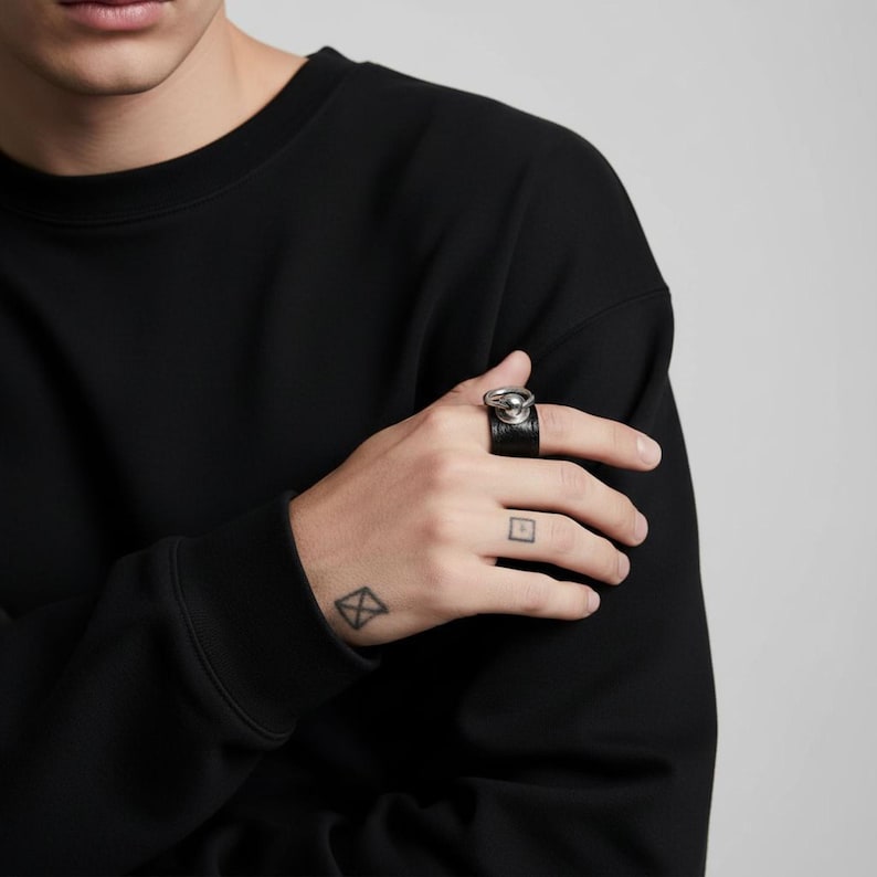 Person wearing black vegan leather ring with polished silver sphere and loop detail. Hand with geometric tattoos resting on black sleeve, minimalist styling with neutral background highlighting the modern unisex jewelry design.