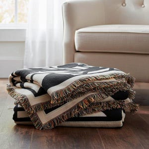 Two folded black and cream geometric woven blankets with multicolor fringed edges stacked on a wooden floor beside a cream upholstered chair. The textured pattern adds a bold graphic touch to a bright, modern interior.