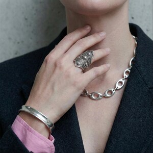Antique silver shield statement ring with ornate floral engraving worn on hand near neck, styled with chunky silver oval chain necklace and polished cuff bracelet. Close-up editorial jewelry styling against concrete background, soft natural light, modern minimalist outfit with dark blazer and pink sleeve detail, bold vintage inspired silver jewelry look, Rannka mood.