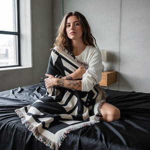Person sitting on a black bed in a minimalist concrete room, wrapped in a black and cream geometric woven blanket with multicolor fringed edges. The blanket features bold angular shapes in a brutalist-inspired pattern. Modern, relaxed, and editorial mood by Rannka.