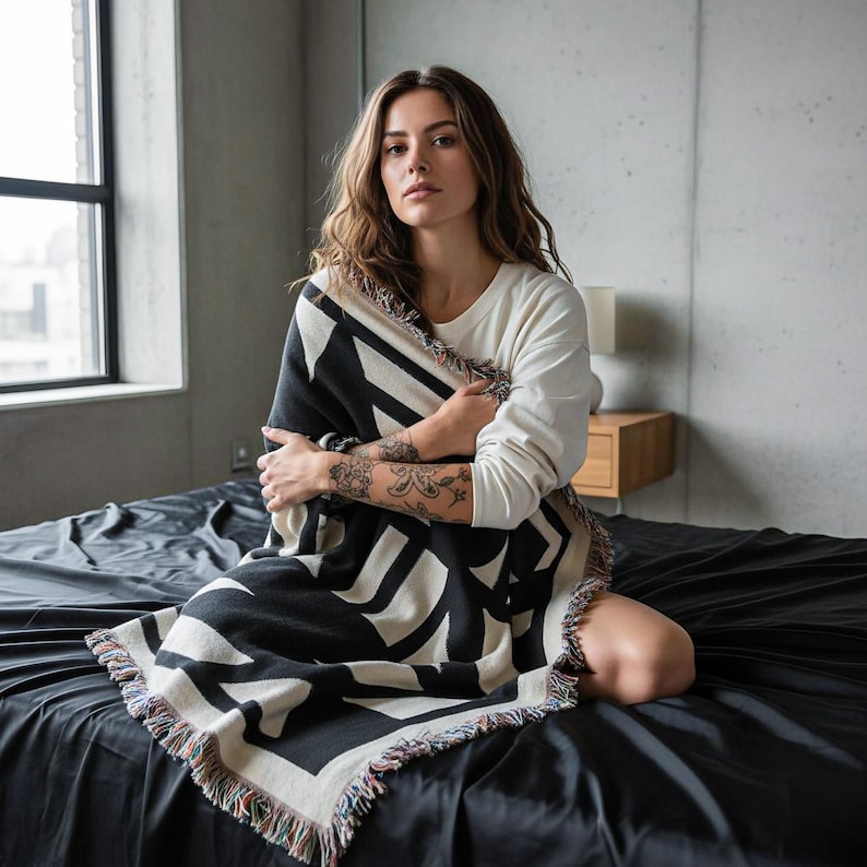 Person sitting on a black bed in a minimalist concrete room, wrapped in a black and cream geometric woven blanket with multicolor fringed edges. The blanket features bold angular shapes in a brutalist-inspired pattern. Modern, relaxed, and editorial mood by Rannka.