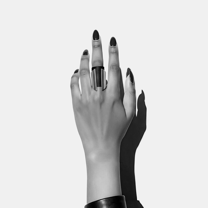 Close-up of a wide black leather and silver-toned ring standing upright on a textured white surface. The design combines soft-grained leather with angled metal panels, creating a sculpted geometric form.