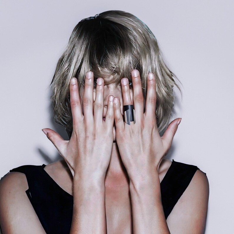 Woman with short blonde hair wearing a sleeveless black top, covering her face with both hands. A wide black leather and silver ring stands out against her skin, emphasizing its sculptural form.