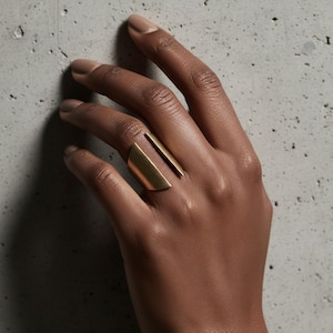Gold sculptural ring with open geometric design worn on hand against a textured concrete background, clean natural lighting highlighting form and simplicity, contemporary minimalist jewelry by Rannka.