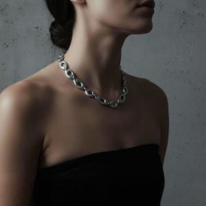 Woman wearing a silver sculptural chain necklace with oval open links, styled with a black strapless top against a textured gray background. The matte metallic finish and fluid form give the piece an architectural, modern edge — minimalist jewelry with strong visual presence.