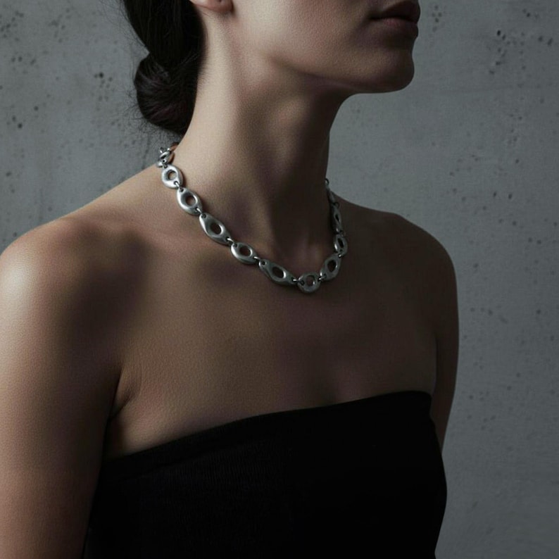 Woman wearing a silver sculptural chain necklace with oval open links, styled with a black strapless top against a textured gray background. The matte metallic finish and fluid form give the piece an architectural, modern edge — minimalist jewelry with strong visual presence.
