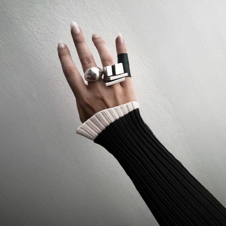 Hand adorned with three sculptural rings — a bold silver dome ring, a geometric bar ring, and a matte black band. Styled with a ribbed black sleeve and pleated cuff. Minimalist, modern, and art-inspired jewelry by Rannka.